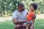 Cute african american boy sitting on fathers knee in park