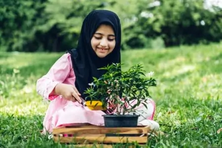 Arabic girl in black hijab sitting on grass planting flowers in a pot