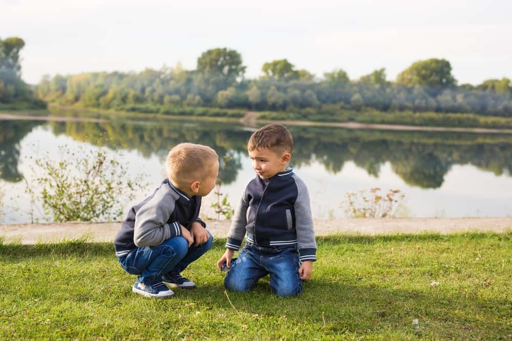 Two european young boys wearing the same jacket sitting on the grass while looking at each other with beautiful nature background