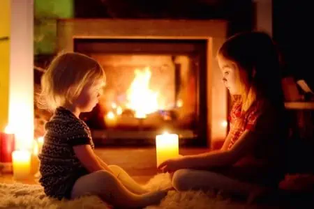 Two adorable little sisters sitting by a fireplace holding candles in a cozy dark living room