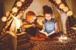 Adorable boy and girl reading book with flashlight in tent at night