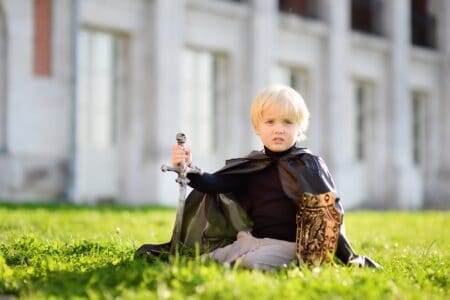 Cute little boy dressed as medieval knight sitting on the grass outdoors