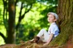 Cute boy wearing hat sitting under an old tree, in the forest