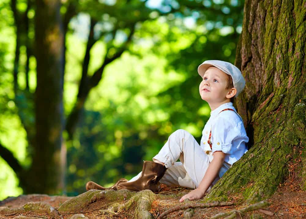 Cute boy wearing hat sitting under an old tree, in the forest