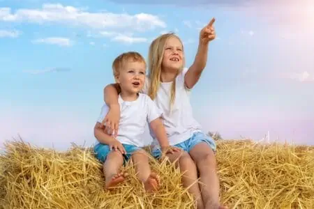 Two cute adorable german siblings having fun while sitting on top over golden hay bale on wheat harvested field near farm