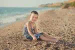 Adorable little boy with exotic charms sitting on the sand near the beach