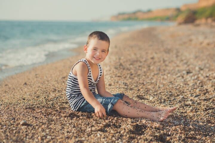 Adorable little boy with exotic charms sitting on the sand near the beach