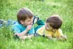 Two boys brothers with magnifying glass outdoors lying on green grass at the park