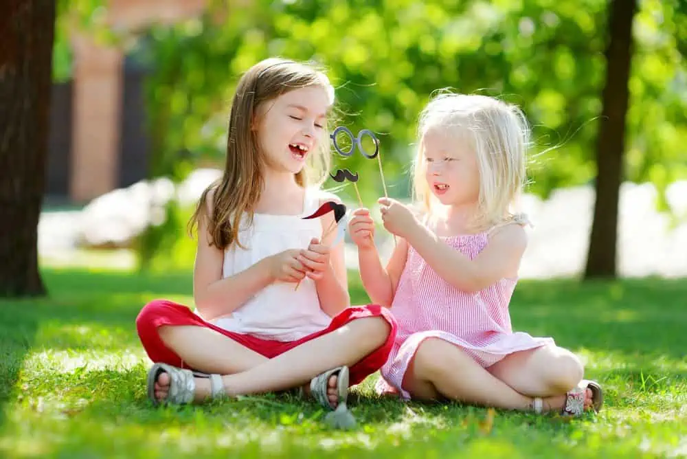 Cheerful little girls playing with paper moustaches on stick at the park