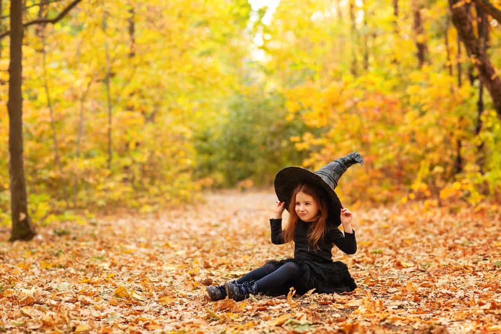 Little toddler girl in witch costume sitting on fallen leaves in autumn park