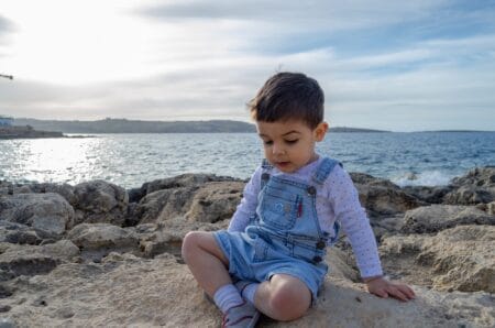 Adorable toddler boy wearing denim jumper and white long sleeves sitting on the rocks by the sea