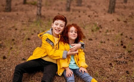 Celtic redhead boy in yellow raincoat embracing sister with curly hair while sitting on grass on autumn day in forest