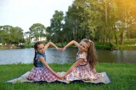 Two little girls sitting on the lawn at the park forming a heart with their hands