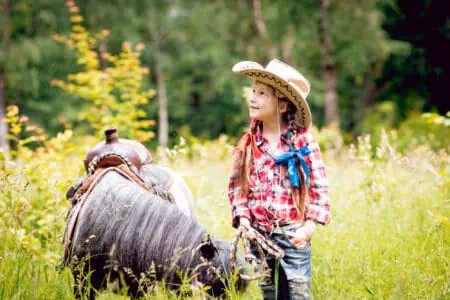 Beautiful little girl wearing cowboy hat standing beside a pony in the meadow