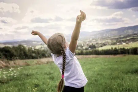 Little girl in pigtails stretching and raising her hands up in the air on the top of the mountains