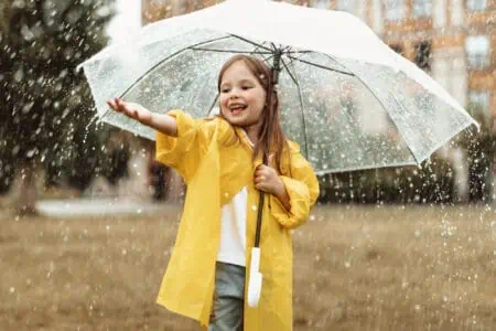 Happy girl in yellow raincoat under umbrella catching raindrops outside