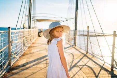Little blonde girl in white dress and a summer hat standing at the pier