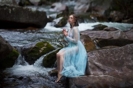 Mythical girl in blue dress sitting on the rock by the river